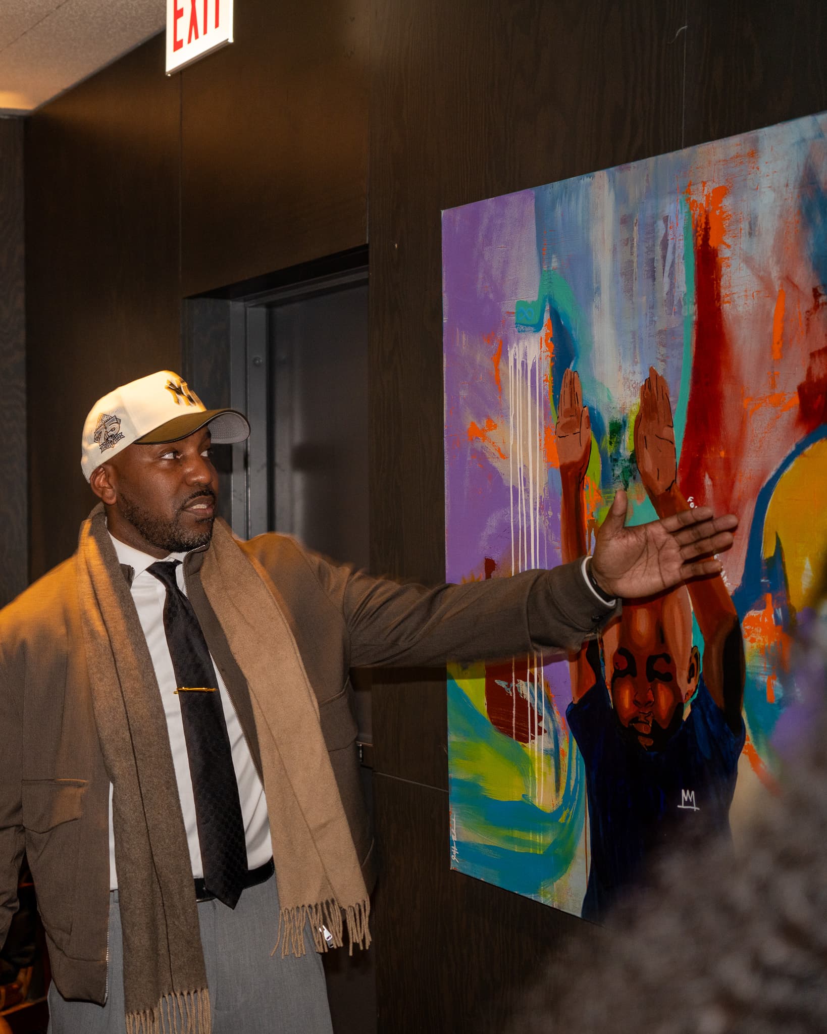 Man in a cream cap and scarf gesturing toward a colorful painting at Jeff Art Hotel