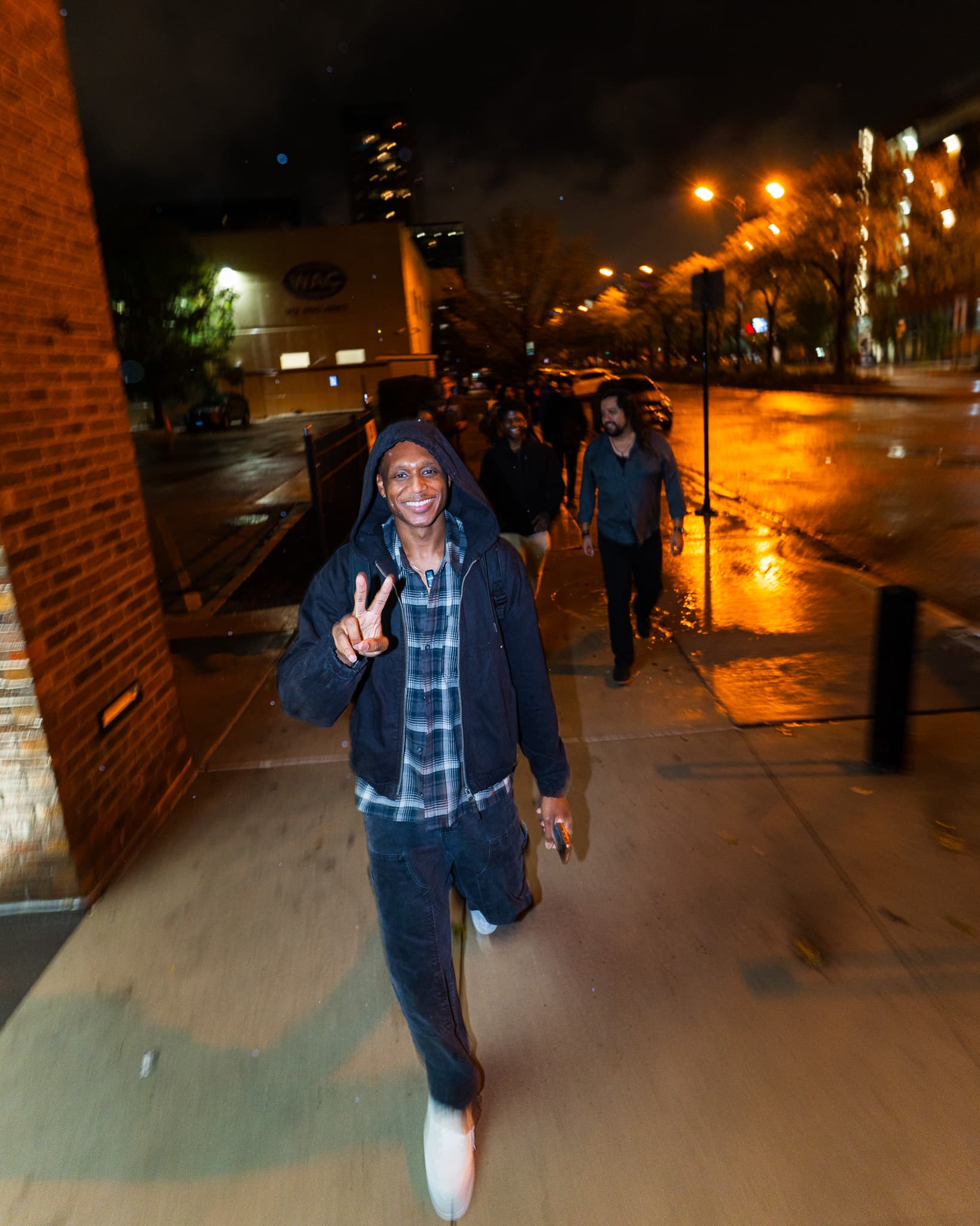 Smiling man in a hoodie throwing a peace sign on a rain-wet sidewalk at night