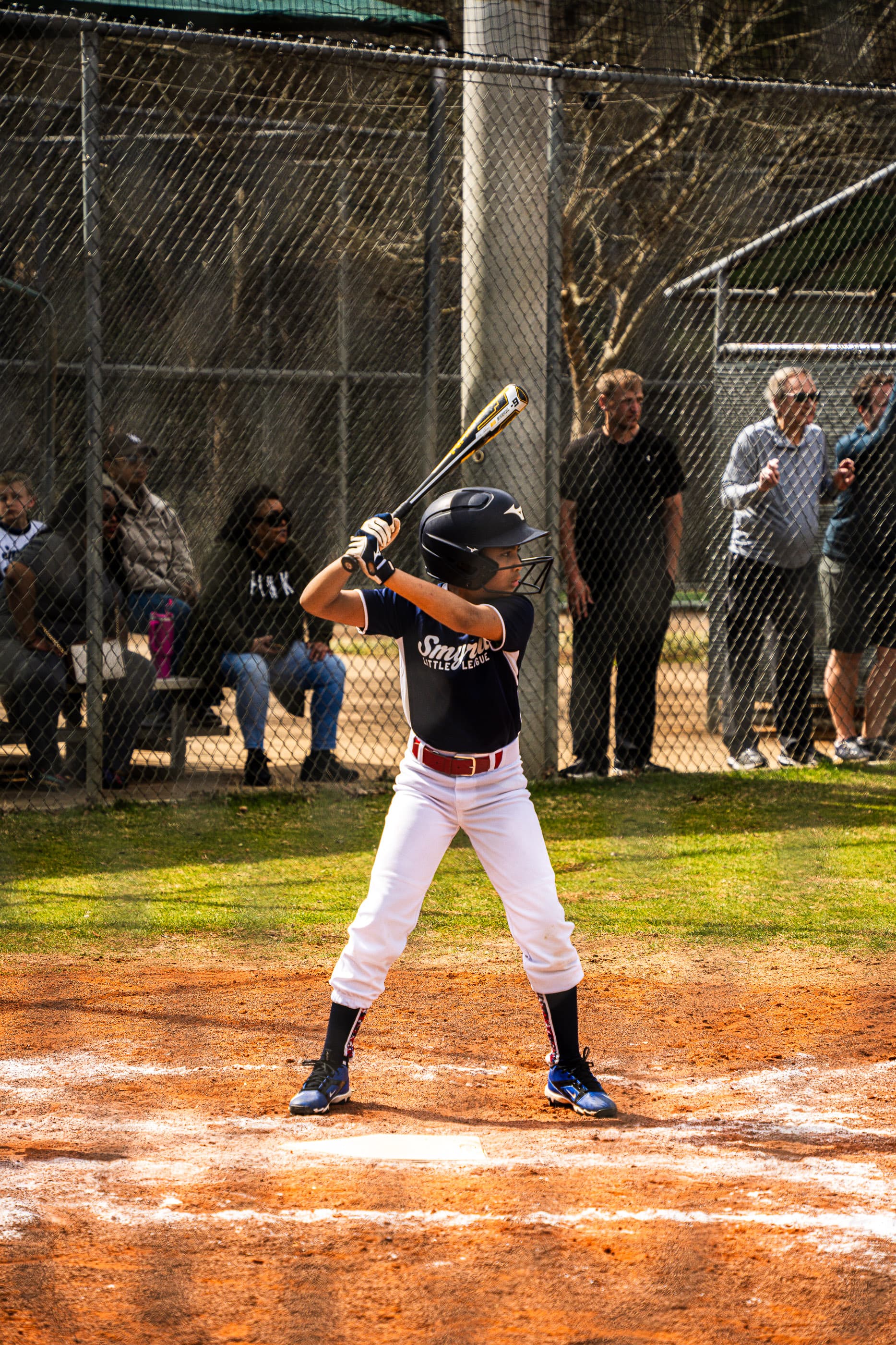 Young batter in a black jersey waiting for the pitch at home plate, spectators behind the fence