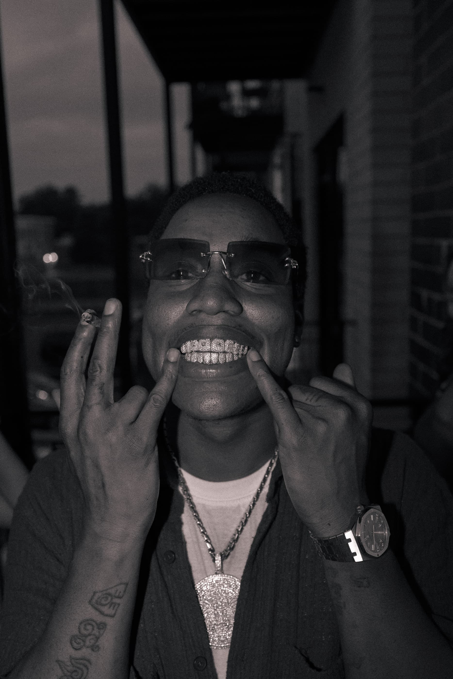 Black and white portrait of a person in sunglasses grinning, hands raised to their face