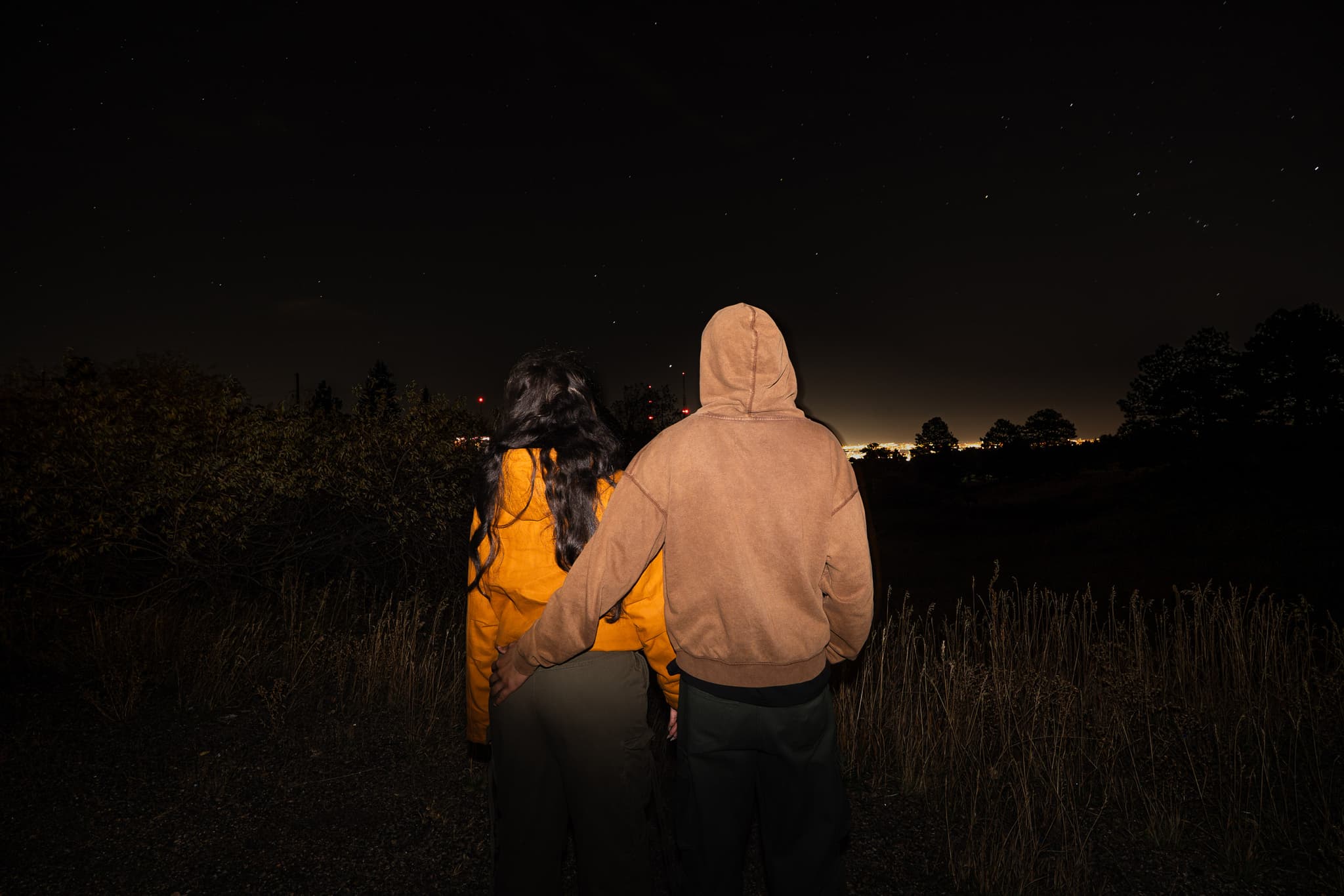 Two people in hoodies standing together in a dark field at night, seen from behind