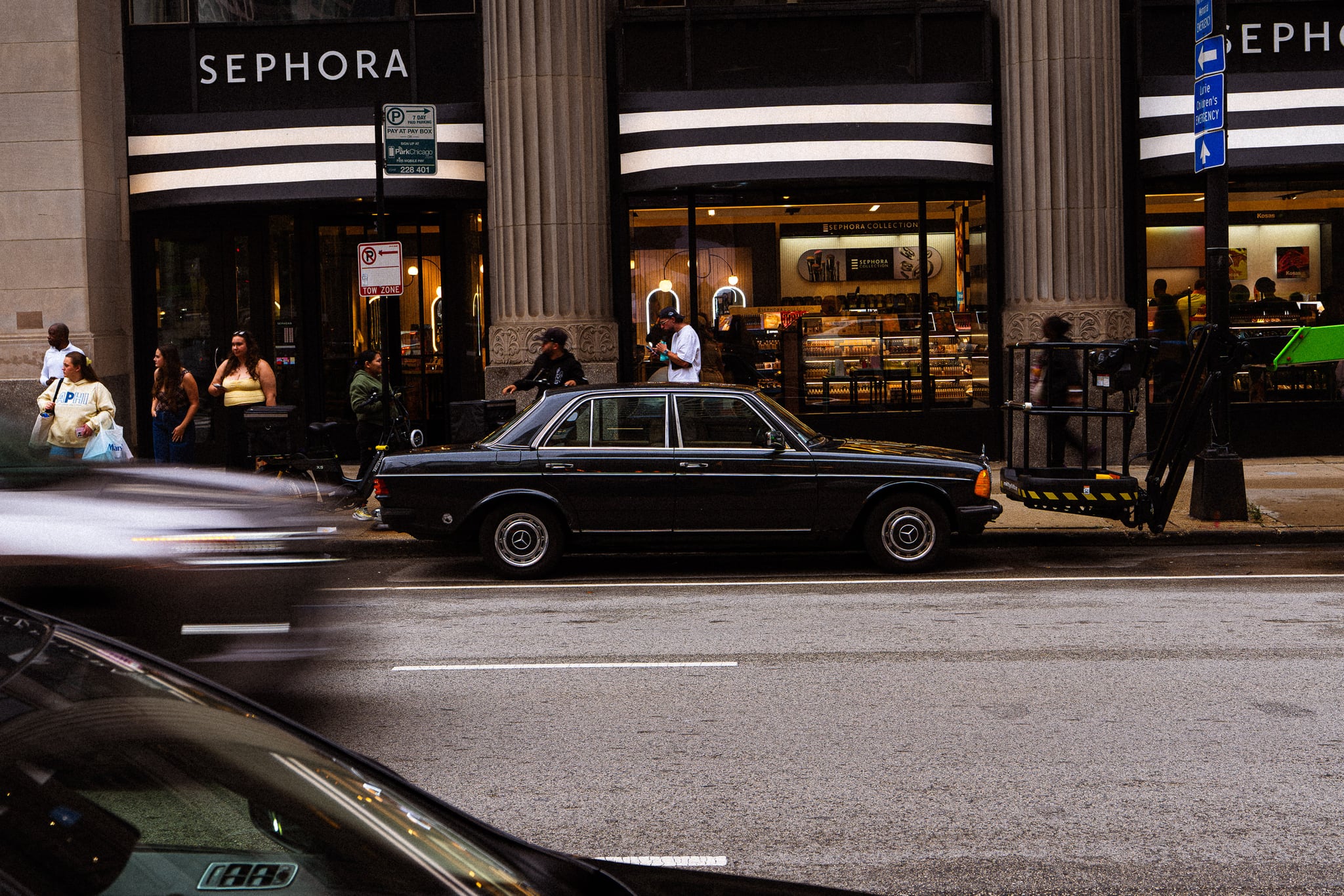 A vintage black sedan parked on a busy shopping street as traffic blurs past
